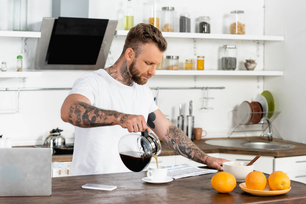 joven hombre tatuado en camiseta blanca leyendo el periódico y vertiendo café cerca de la computadora portátil y teléfono inteligente en la mesa de la cocina - Foto, Imagen