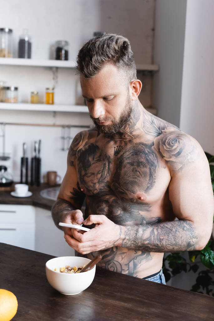 τατουάζ, μυώδης άνθρωπος χρησιμοποιώντας φορητό υπολογιστή, ενώ στέκεται κοντά μπολ με πρωινό στην κουζίνα - Φωτογραφία, εικόνα