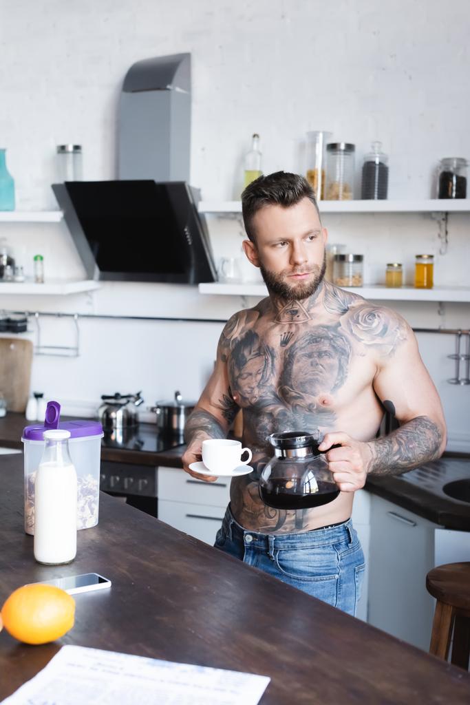 Hemdlos tätowierter Mann schaut weg, während er mit Kaffeekanne und Tasse in der Küche steht - Foto, Bild