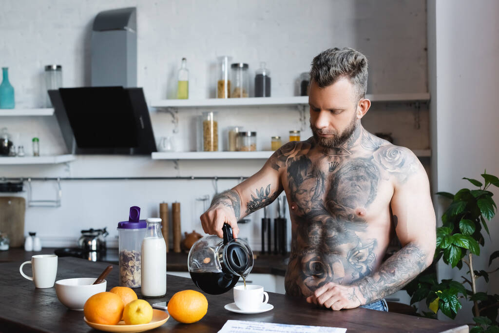sin camisa, hombre tatuado vertiendo café en la taza durante el desayuno en la cocina - Foto, Imagen