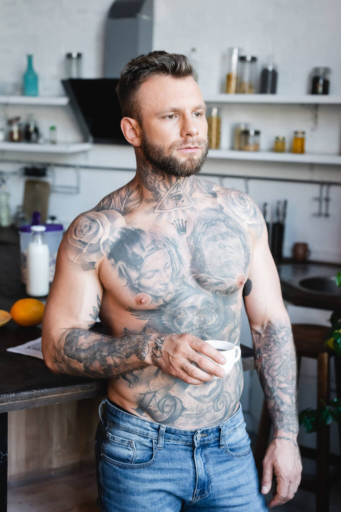 fiatal, félmeztelen tetovált férfi félrenéz, miközben egy csésze kávét tart a konyhában. - Fotó, kép