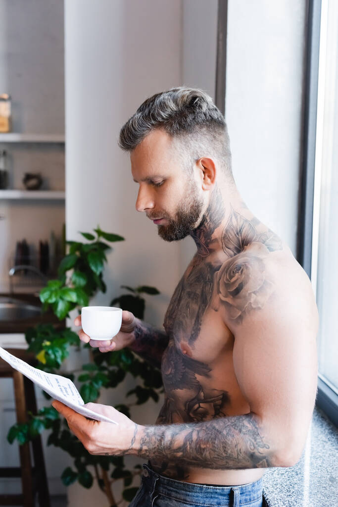 puolella näkymä paidaton tatuoitu mies lukee sanomalehteä pitäessään kahvikupin keittiössä - Valokuva, kuva