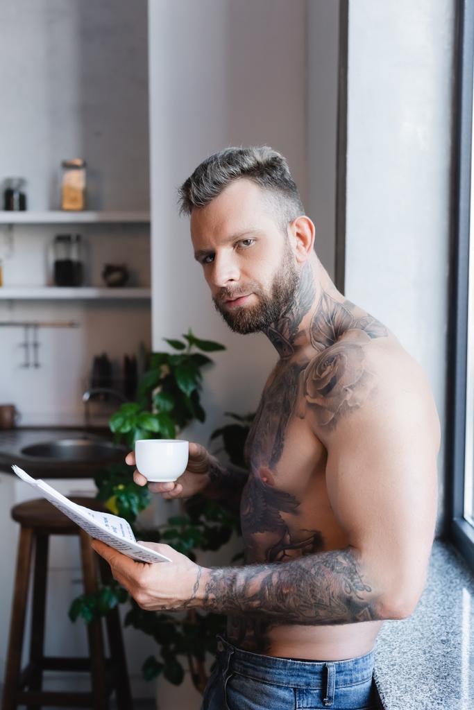 joven hombre tatuado sosteniendo taza de café y periódico durante el desayuno en la cocina - Foto, Imagen