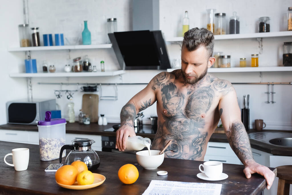 hombre tatuado sin camisa verter leche en un tazón cerca de naranjas frescas y periódico durante el desayuno en la cocina - Foto, imagen