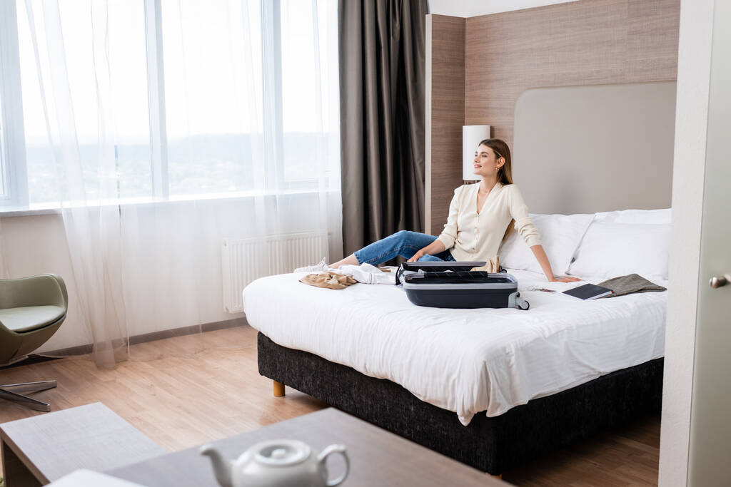 selective focus of young woman looking away near notebook and luggage on bed in hotel - Photo, Image