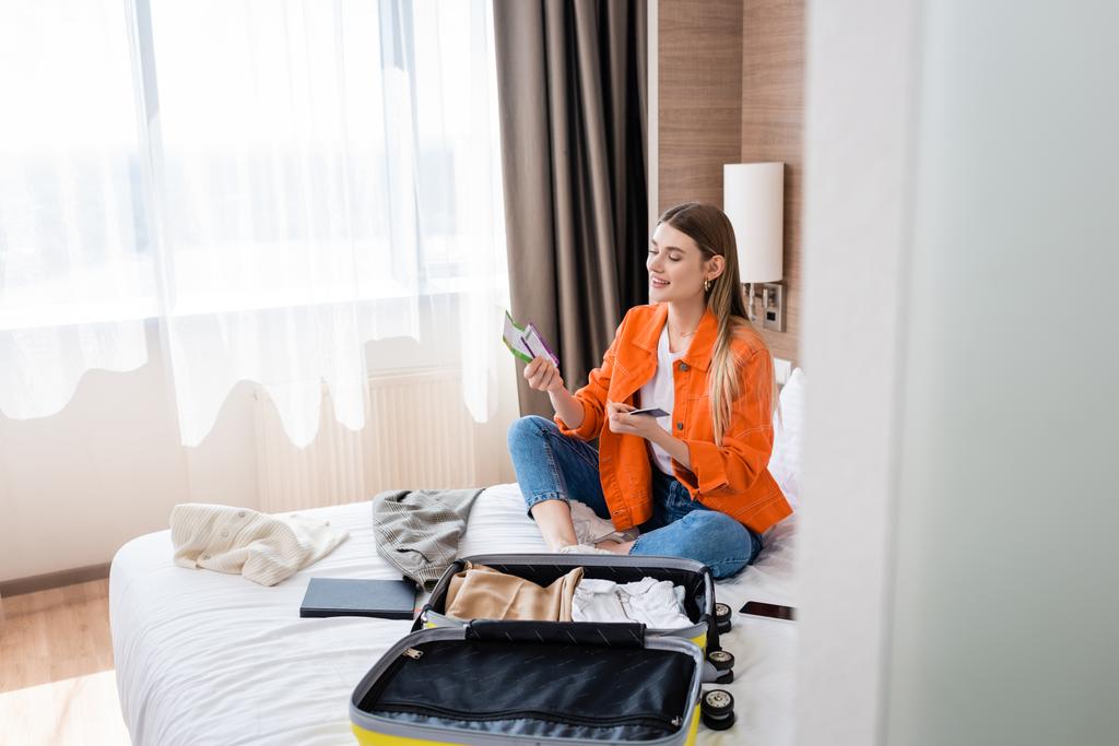 Selektiver Fokus der jungen Frau mit Pass, Bordkarte und Flugticket in der Nähe von Gepäck und Notizbuch im Hotelbett  - Foto, Bild