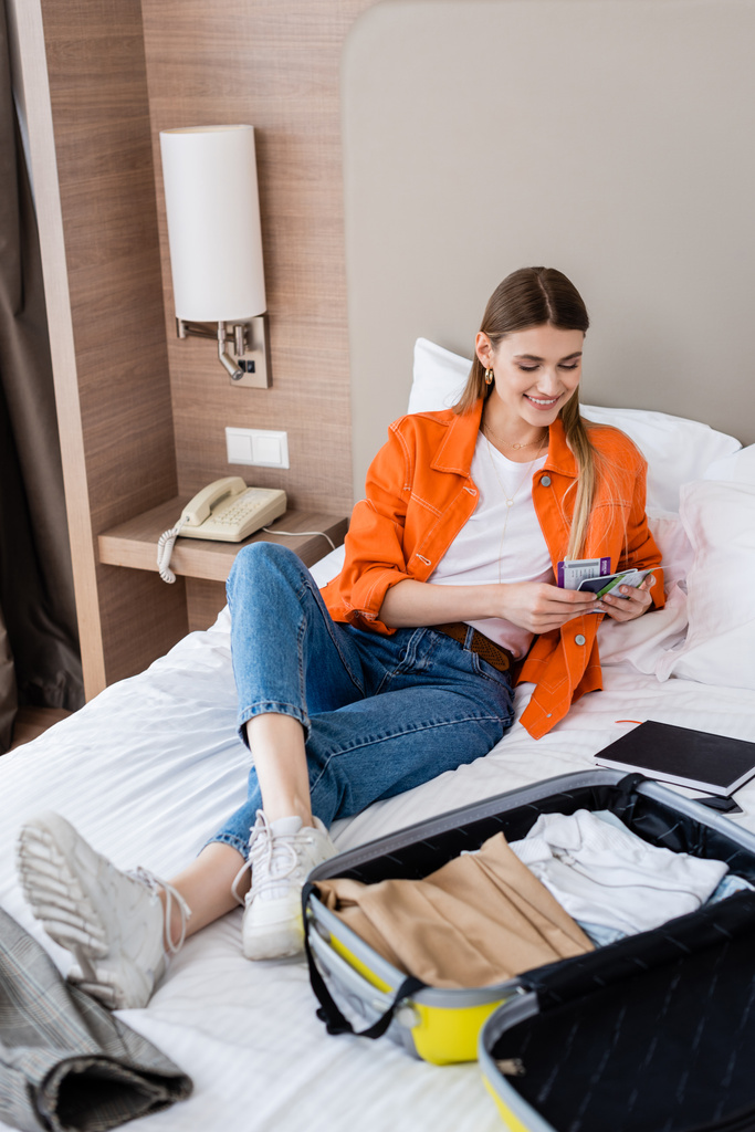 femme joyeuse tenant passeport, carte d'embarquement et billet d'avion près des bagages et carnet sur le lit dans l'hôtel  - Photo, image