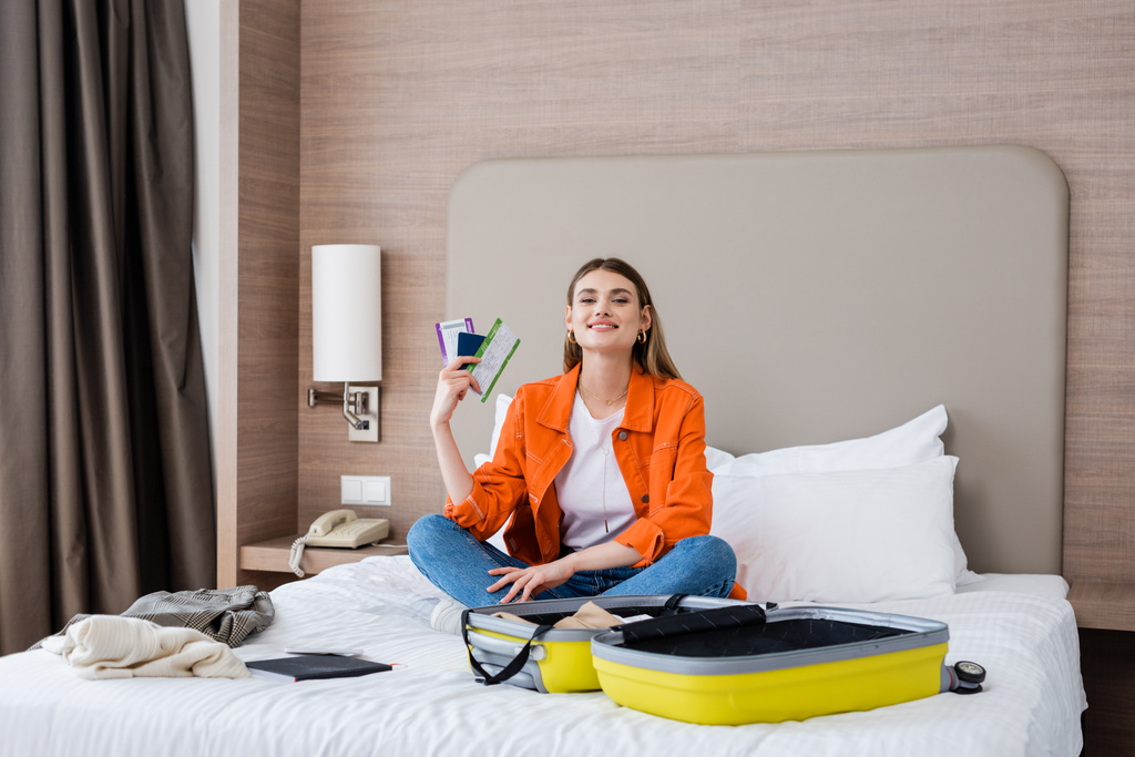 pleased woman holding passport, boarding pass and air ticket near baggage and notebook on bed in hotel  - Photo, Image