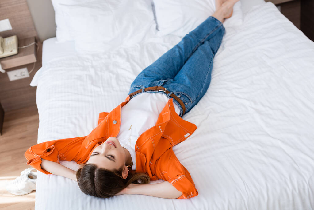άποψη υψηλής γωνίας της νεαρής γυναίκας σε τζιν παντελόνι στηρίζεται στο κρεβάτι στο ξενοδοχείο - Φωτογραφία, εικόνα