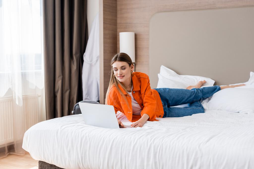 裸足の女性がベッドに寝そべってホテルの部屋でノートパソコンを使って - 写真・画像