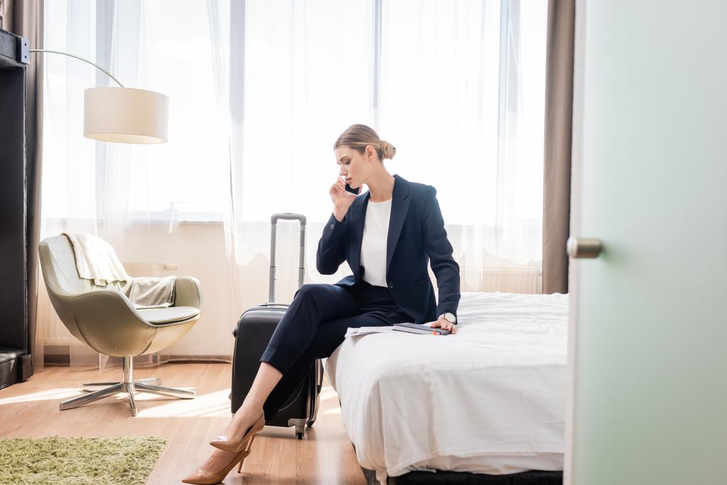 wybiórcze skupienie bizneswoman w garniturze mówi na smartfonie siedząc na łóżku w pobliżu bagażu w hotelu  - Zdjęcie, obraz
