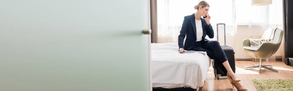 vízszintes kép üzletasszony öltönyben beszél okostelefonon, miközben ül az ágyon közelében poggyász a szállodában  - Fotó, kép