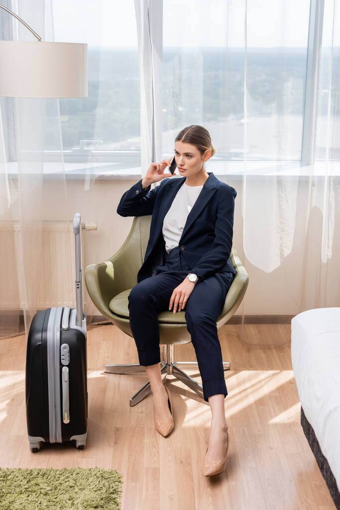 młoda bizneswoman w garniturze rozmawia na smartfonie i siedzi w fotelu w pobliżu bagażu w hotelu  - Zdjęcie, obraz