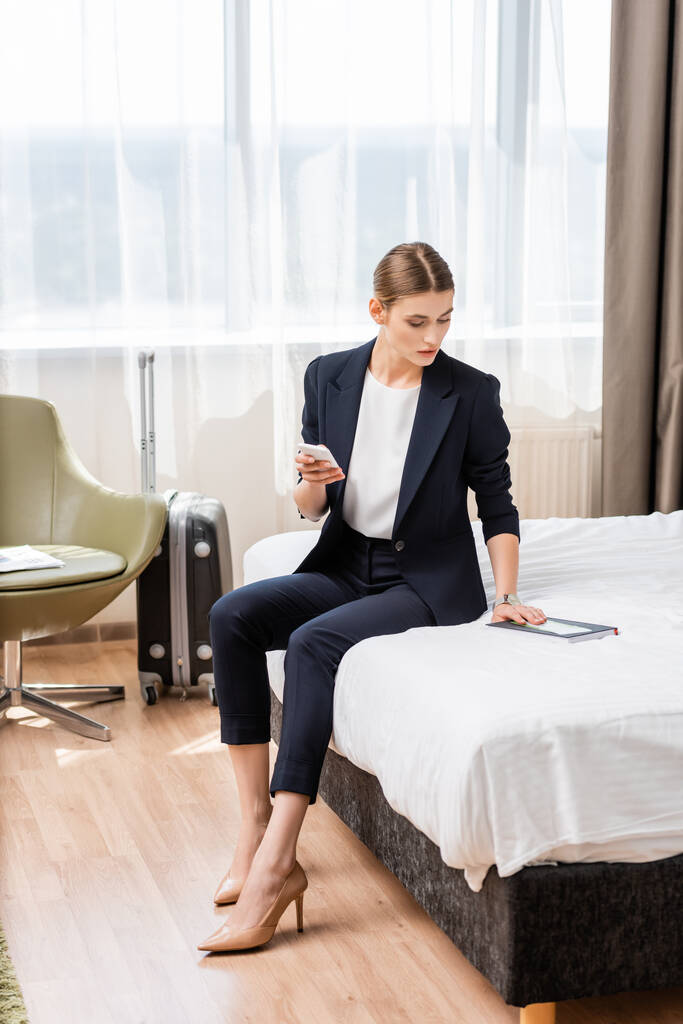 üzletasszony öltönyben gazdaság okostelefon és nézi notebook repülőjegy közelében poggyász a szállodai szobában  - Fotó, kép