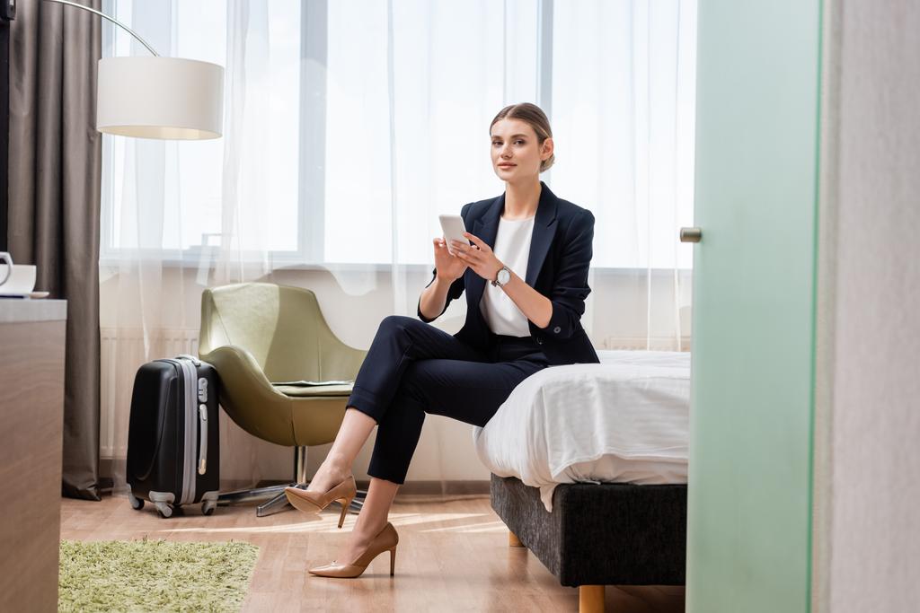 Takım elbiseli genç bir iş kadını elinde akıllı bir telefonla otel odasında temiz bir şekilde seyahat çantasında oturuyor.  - Fotoğraf, Görsel