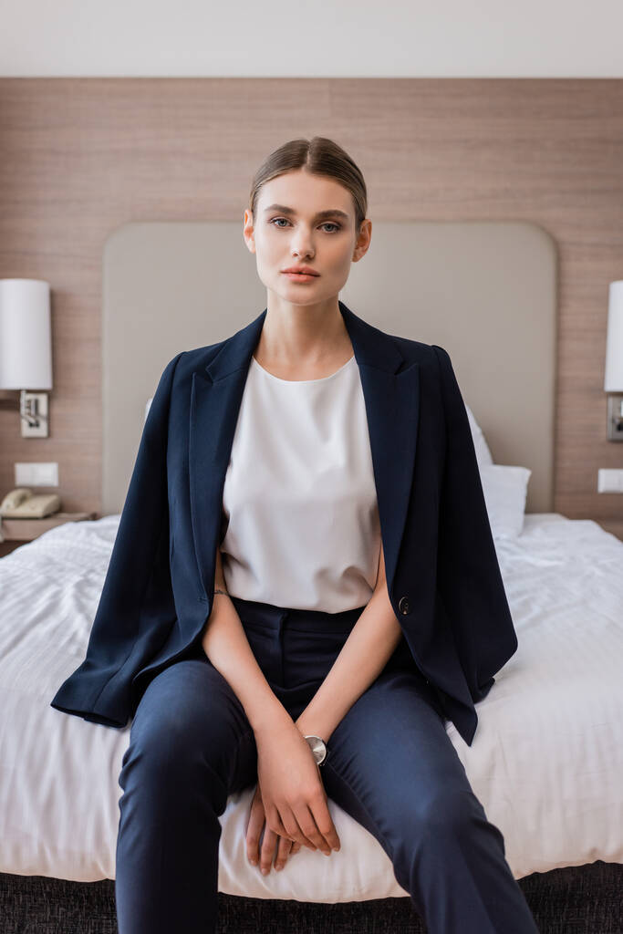 Geschäftsfrau in offizieller Kleidung blickt in die Kamera und sitzt im Hotel auf dem Bett  - Foto, Bild