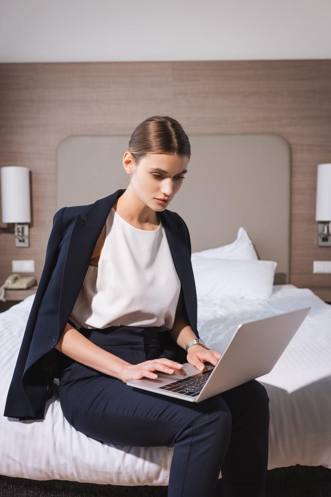 mujer en traje sentado en la cama y el uso de la computadora portátil en la habitación de hotel  - Foto, Imagen
