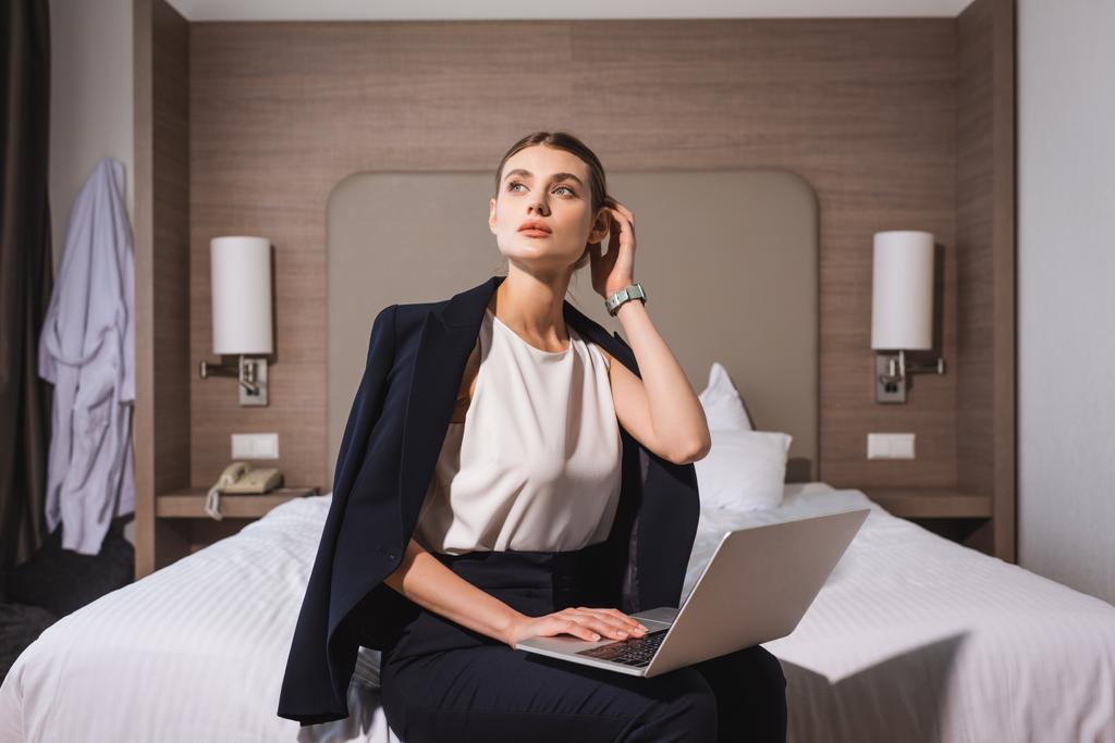 γυναίκα με κοστούμι κάθεται στο κρεβάτι με φορητό υπολογιστή και αγγίζοντας τα μαλλιά στο δωμάτιο του ξενοδοχείου  - Φωτογραφία, εικόνα