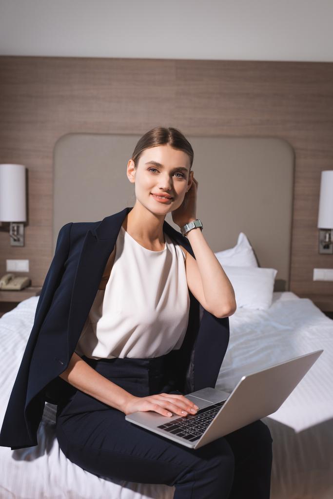 ευχαριστημένη γυναίκα με κοστούμι που κάθεται στο κρεβάτι με φορητό υπολογιστή στο δωμάτιο του ξενοδοχείου  - Φωτογραφία, εικόνα