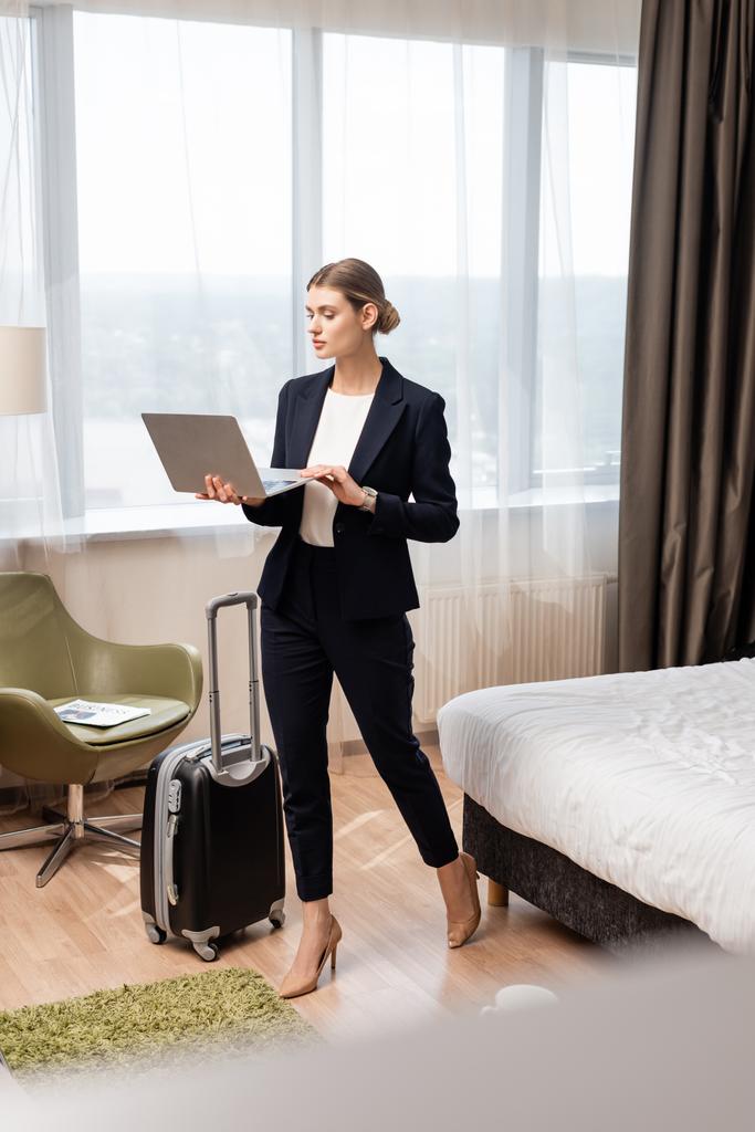 joven mujer de negocios de pie y utilizando el ordenador portátil cerca de la bolsa de viaje en la habitación de hotel  - Foto, imagen
