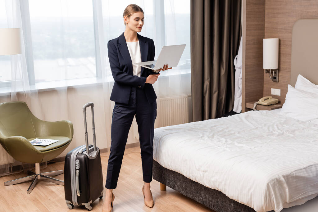 bizneswoman stojąca i korzystająca z laptopa w pobliżu torby podróżnej w pokoju hotelowym  - Zdjęcie, obraz