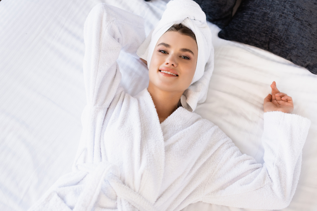 von oben freudige Frau in Handtuch und weißem Bademantel auf Bett im Hotelzimmer liegend - Foto, Bild