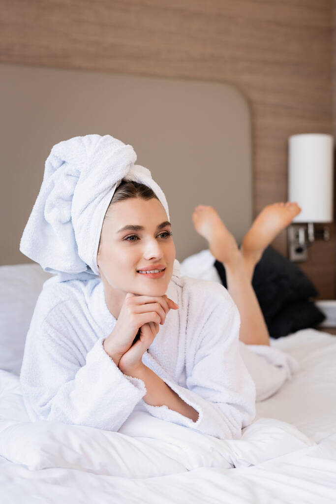 mujer descalza en toalla y albornoz blanco acostada en la cama y mirando hacia otro lado en la habitación del hotel  - Foto, imagen