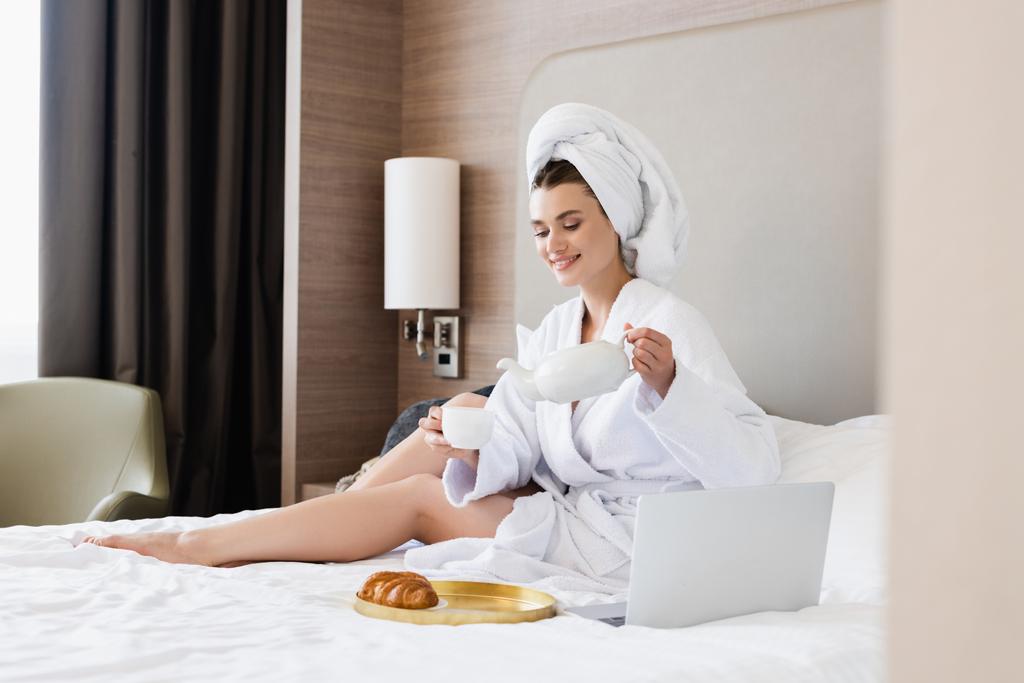 donna in accappatoio in possesso di tazza e teiera vicino al computer portatile e vassoio con colazione sul letto - Foto, immagini