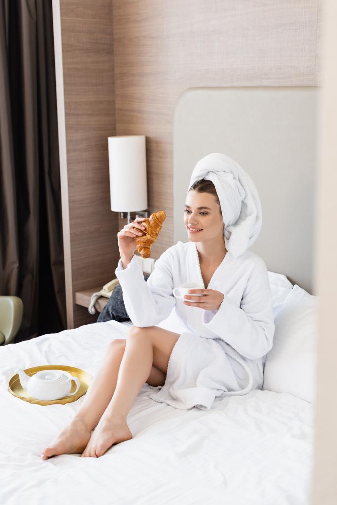 barefoot vrouw in handdoek en badjas houden croissant en kopje koffie in de buurt van ontbijt lade op bed  - Foto, afbeelding