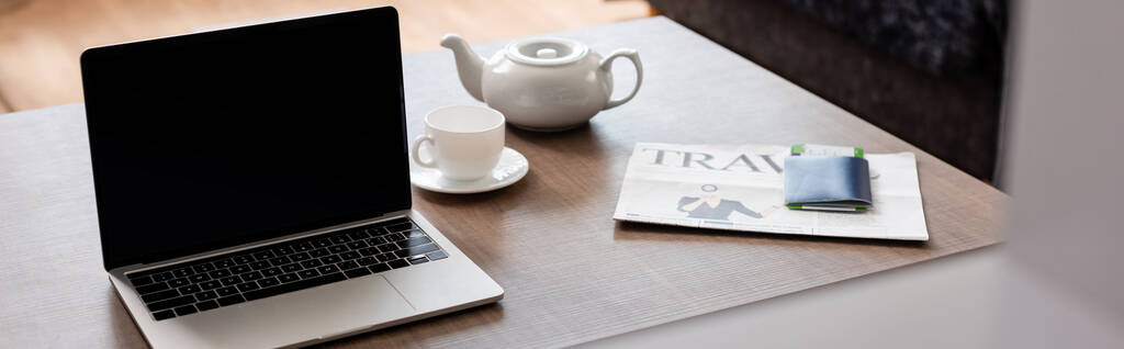 panorámás termés laptop üres képernyővel közel teáskanna, csésze, utazási újság és útlevél repülőjegy az asztalon  - Fotó, kép