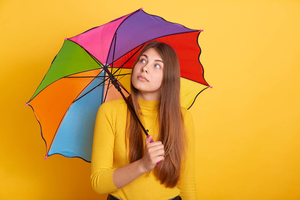 Pensive ελκυστική γυναίκα κρατώντας πολύχρωμη ομπρέλα και κοιτάζοντας στην άκρη, κορίτσι με μακριά όμορφα μαλλιά, στέκεται ενάντια στον κίτρινο τοίχο σε casual ρούχα. - Φωτογραφία, εικόνα