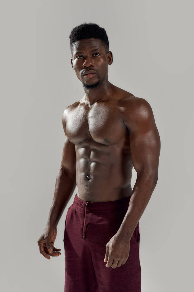 Υγιές σώμα. Μυώδης Αφροαμερικάνος που κοιτάζει την κάμερα, δείχνοντας τον γυμνό κορμό του ενώ παριστάνει τον γυμνό απομονωμένο πάνω σε γκρι φόντο. Αθλητισμός, προπόνηση, bodybuilding έννοια - Φωτογραφία, εικόνα
