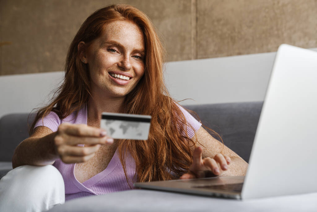 Зображення рудої усміхненої жінки, що тримає кредитну картку і використовує ноутбук, сидячи на підлозі вдома
 - Фото, зображення