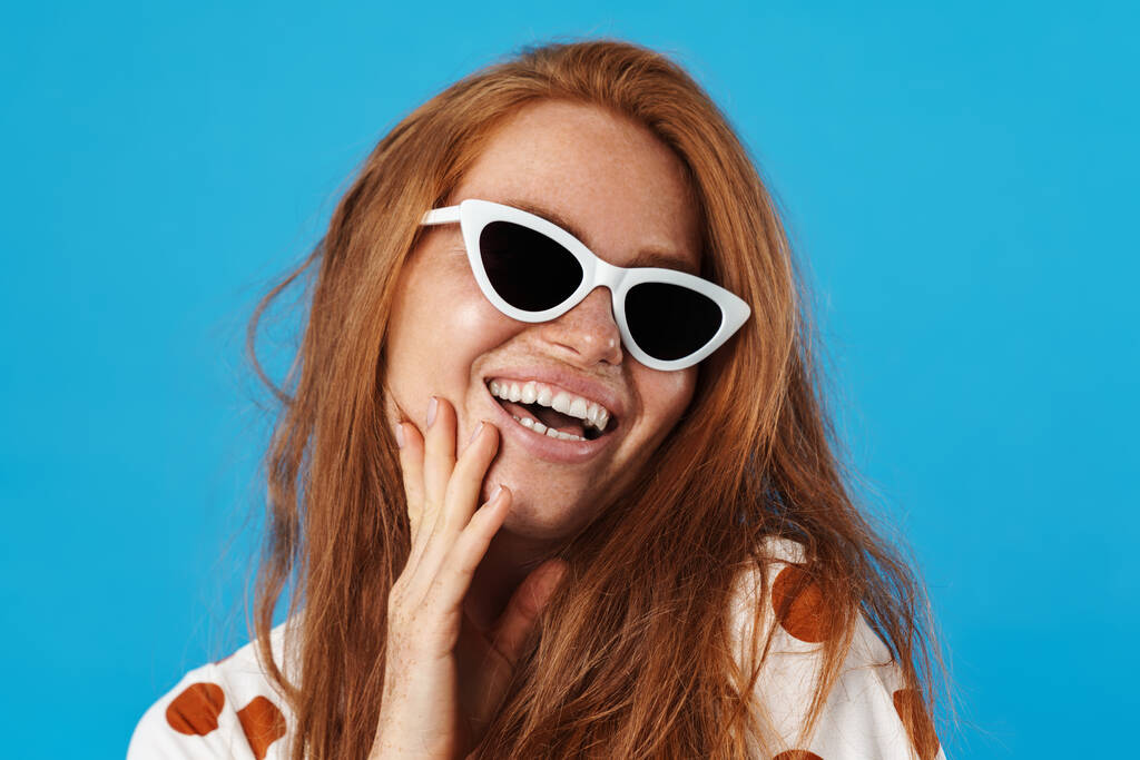 Portrait of a beautiful cheerful young woman with long red hair wearing summer clothes and sunglasses standing isolated over blue background, posing - Photo, Image