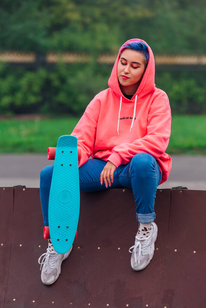 Giovane ragazza con capelli corti multicolore siede con skateboard in skatepark - Foto, immagini
