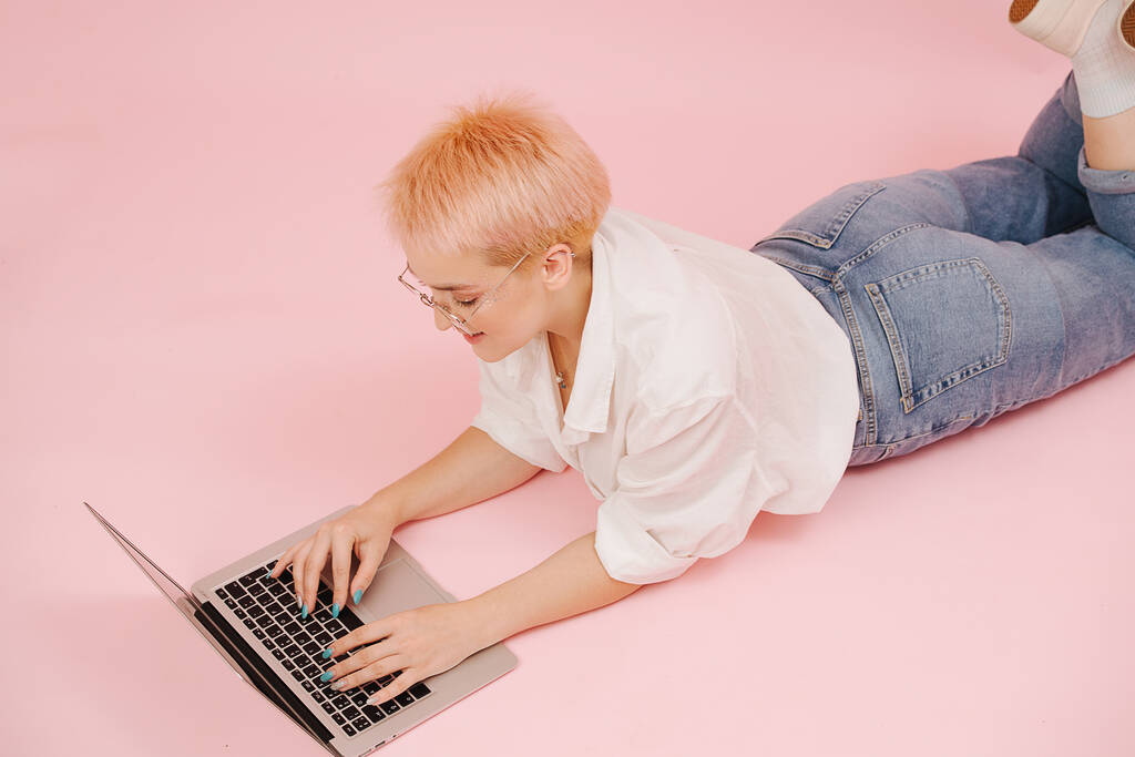 Eine junge Frau mit kurz gefärbten blonden Haaren liegt auf dem Boden und arbeitet an einem Laptop. Sie trägt Mama-Jeans und Kleid-Hemd. Über rosa Wand mit grünen und gelben Papierformen, die darauf geklebt sind. - Foto, Bild