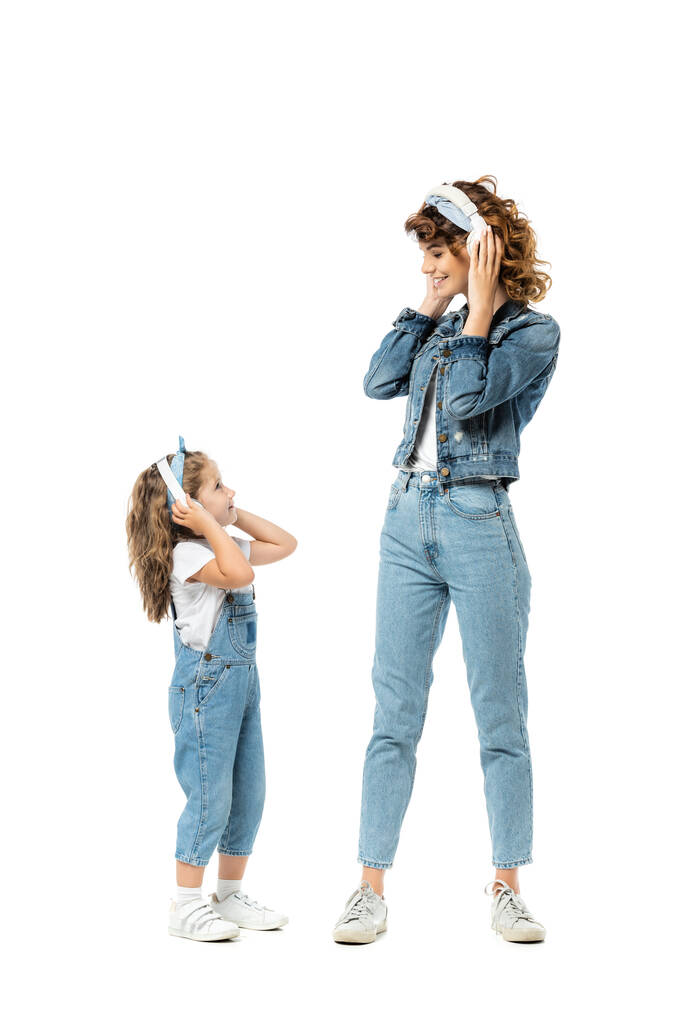 Mutter und Tochter im Jeans-Outfit hören Musik über Kopfhörer und schauen sich isoliert auf weißem Grund an - Foto, Bild