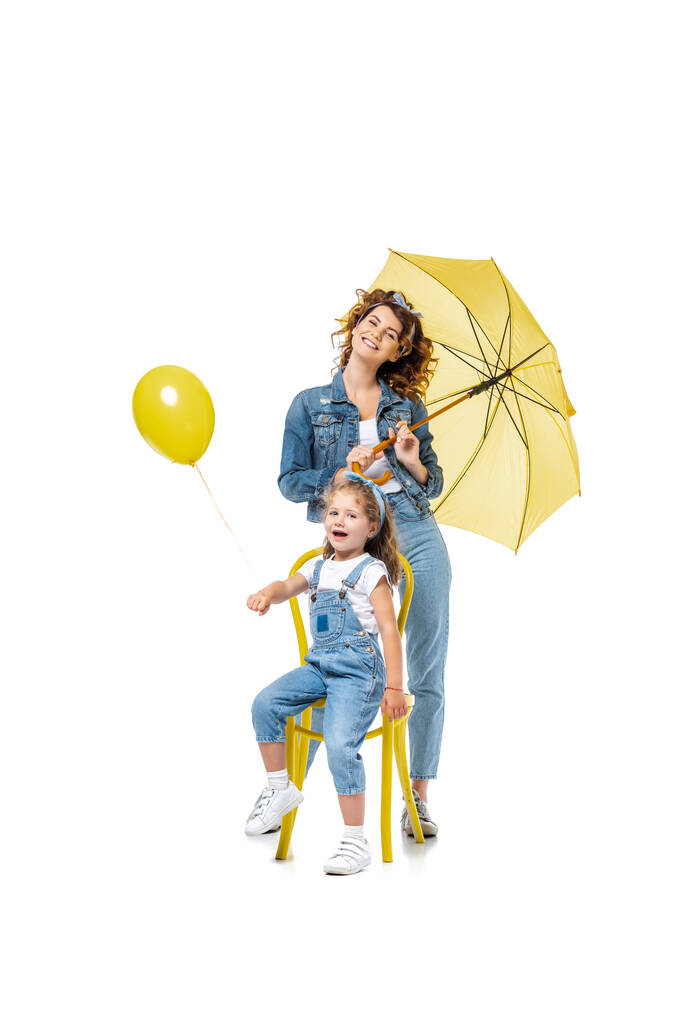 madre con paraguas amarillo de pie cerca de la hija en silla con globo aislado en blanco - Foto, Imagen