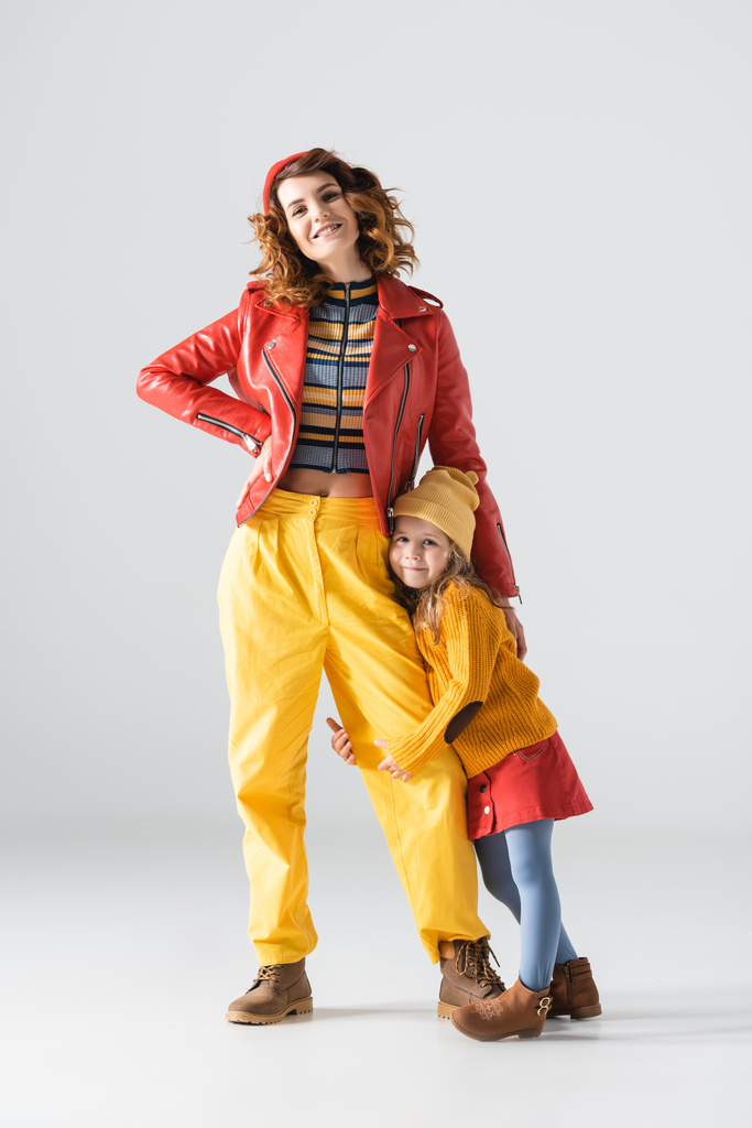 figlia in abito colorato rosso e giallo abbracciando gamba madre su sfondo grigio - Foto, immagini