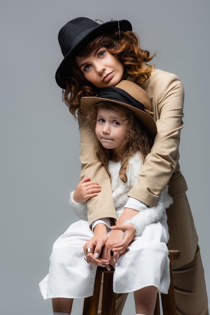 κομψή μητέρα και κόρη σε λευκό και μπεζ ρούχα αγκαλιάζει σε καρέκλα απομονωμένη σε γκρι - Φωτογραφία, εικόνα