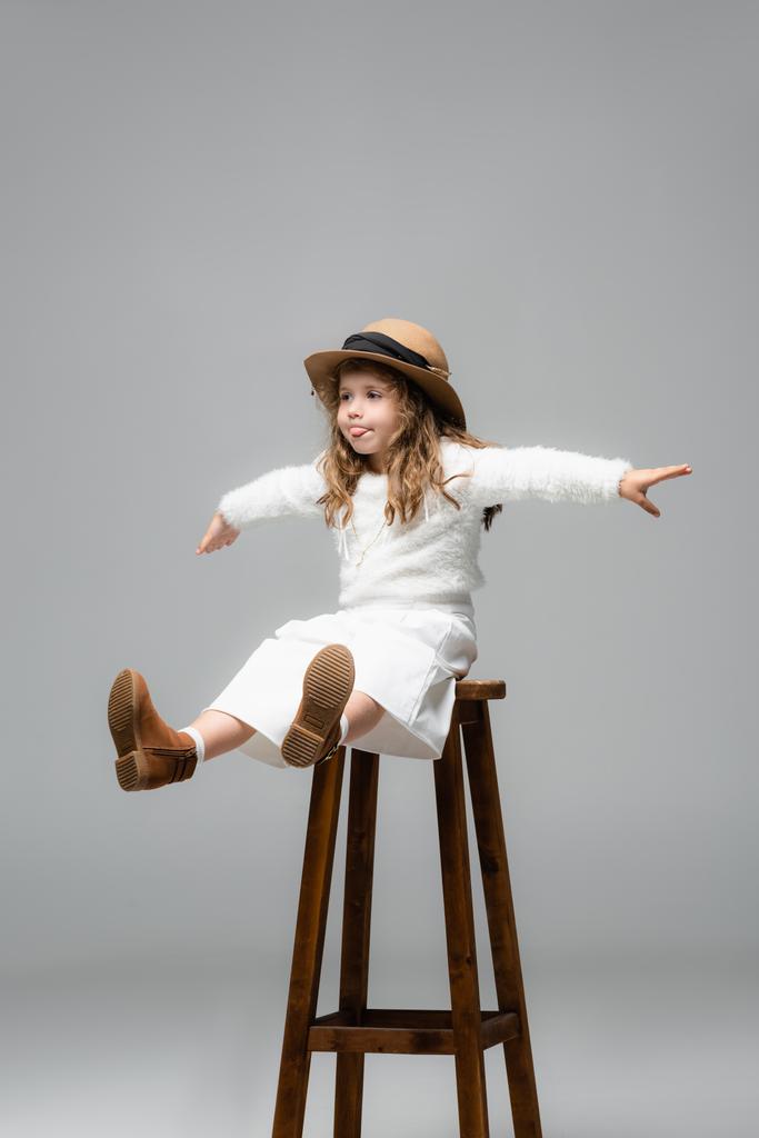 menina elegante em roupa branca posando na cadeira com as mãos estendidas no cinza - Foto, Imagem