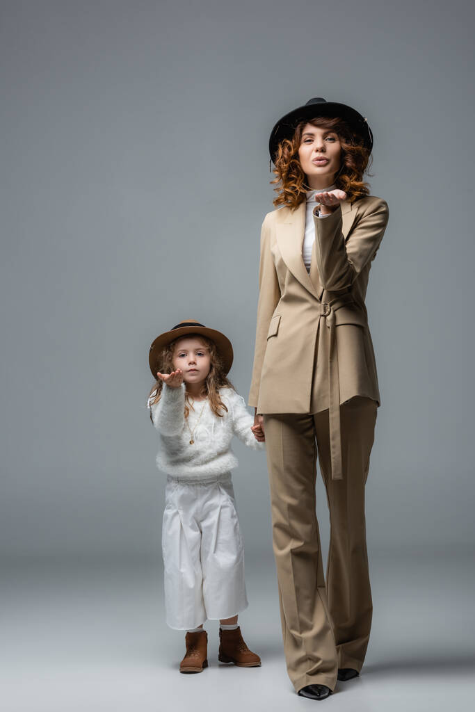 elegante Mutter und Tochter in weiß-beigen Outfits und Hüten, die Küsse blasen und Händchen halten auf grau - Foto, Bild