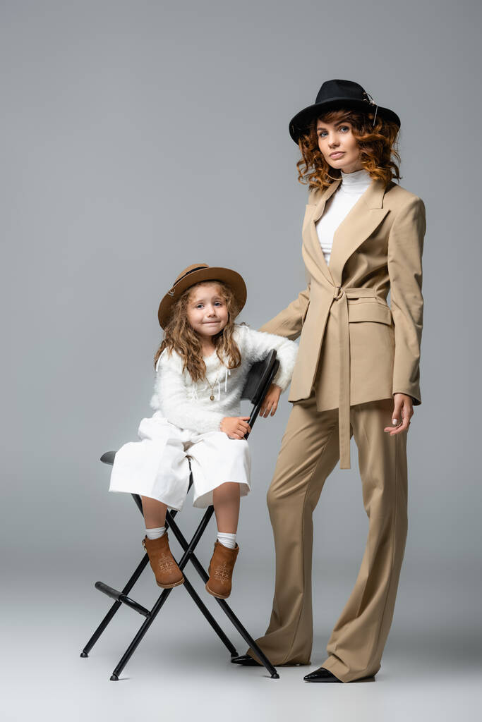 elegancka matka i córka w biało-beżowych strojach i kapeluszach pozujących na krześle na szarym tle - Zdjęcie, obraz