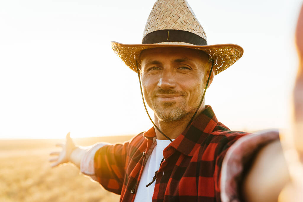 Imagen del hombre adulto complacido en sombrero de paja tomando foto de selfie mientras está de pie en el campo de cereales - Foto, imagen