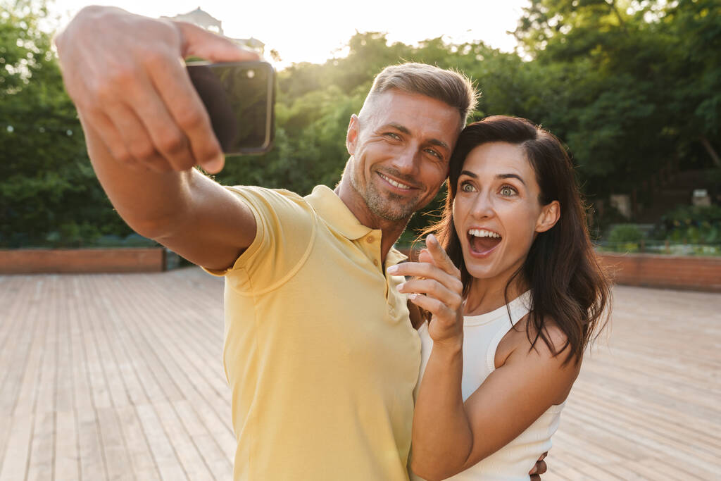 興奮した大人のカップルの男性と女性の肖像写真を携帯電話で撮影し、指を指しながら夏の公園を歩きます - 写真・画像