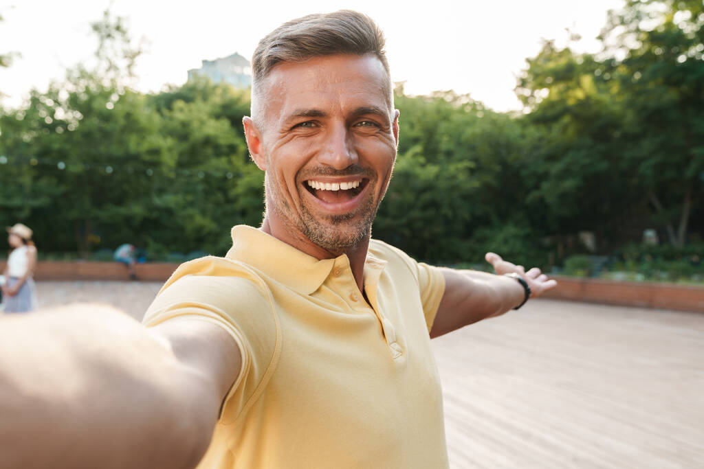 Immagine primo piano di felice uomo di mezza età che indossa t-shirt gialla scattare foto selfie e ridere mentre si cammina nel parco estivo - Foto, immagini
