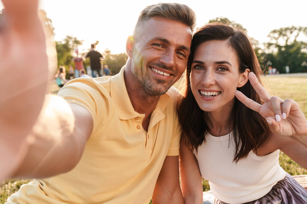 Image d'un couple aimant adulte mignon positif joyeux à l'extérieur dans un beau parc naturel vert prendre un selfie par caméra montrant geste de paix. - Photo, image