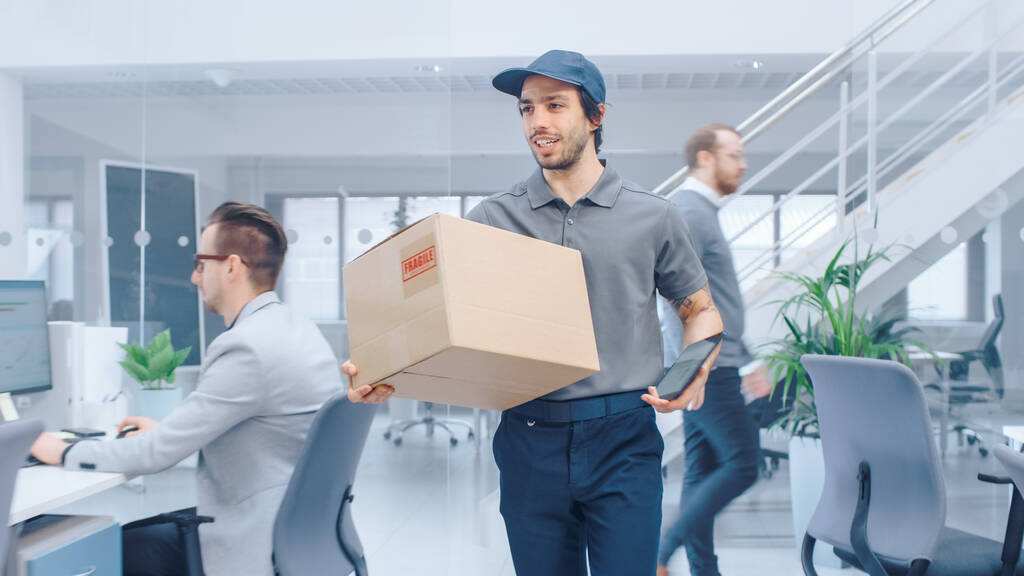 Happy Delivery Man Enters Corporate Office and Delivers Cardboard Package to a Worker. Big Bright Modern Business Company Office with Professional Businesspeople Working - Photo, Image