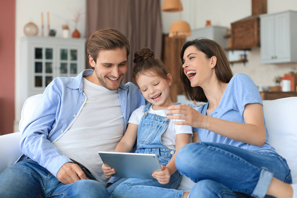 Pozytywni młodzi rodzice z uśmiechniętą córeczką siedzącą razem na kanapie, odpowiadający na wideo połączenie na tablecie cyfrowym, relaksujący się w domu w weekend - Zdjęcie, obraz