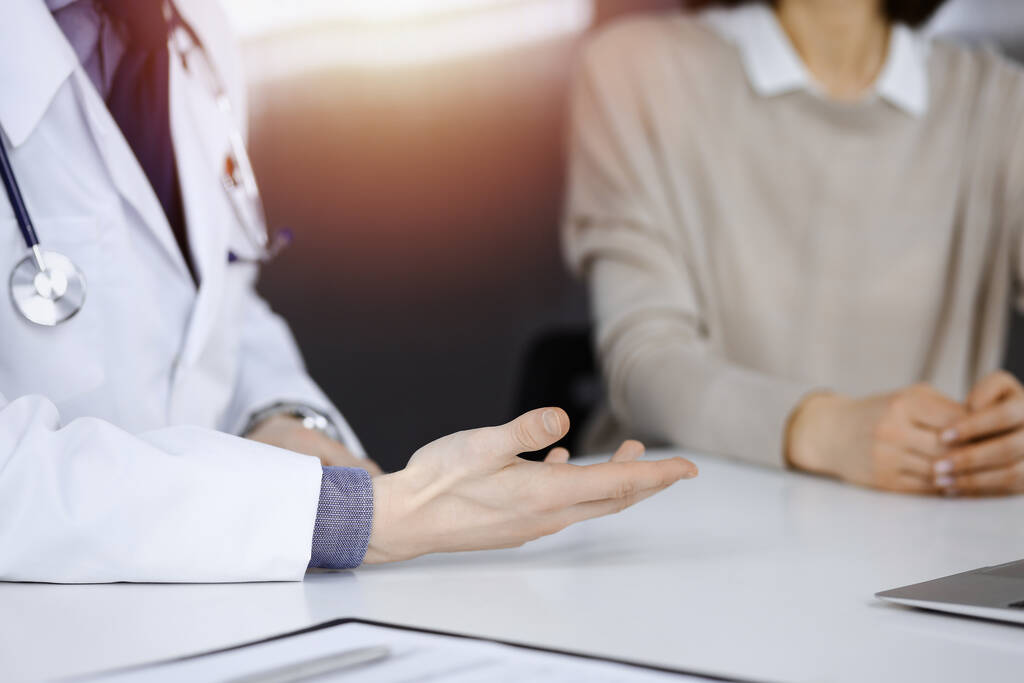 Médico desconhecido e mulher paciente discutindo algo enquanto se senta em uma clínica escurecida, brilho de luz no fundo. Close-up de mãos - Foto, Imagem